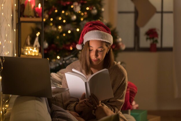 Woman with santa hat reading book in front of laptop
