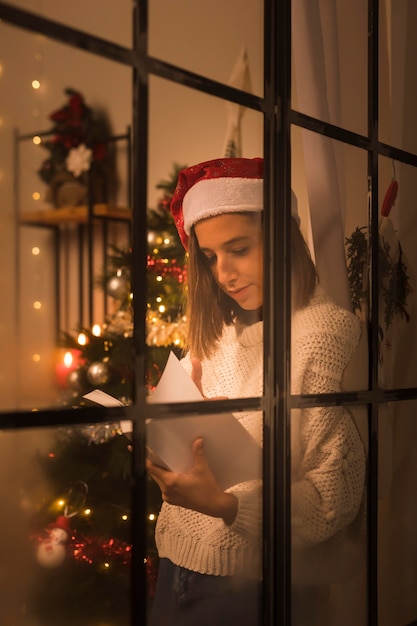 Free photo woman with santa hat through window holding and reading book on christmas