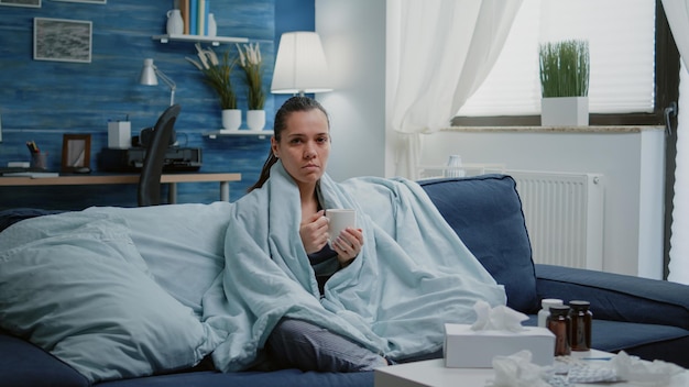 Woman with seasonal cold holding cup of tea wrapped in blanket. Sick adult with flu looking at camera while feeling ill and shivering. Portrait of person with medicaments on table