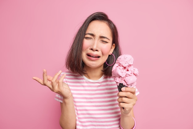 woman with short dark hair cries from despair expresses negative emotions feels displeased holds big delicious gelato wears casual striped t shirt has broken heart