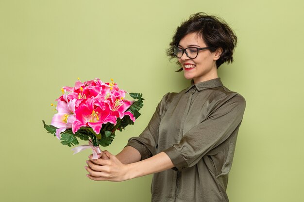 Woman with short hair holding bouquet of flowers looking at flowers happy and pleased smiling cheerfully celebrating international women's day march 8 standing over green background