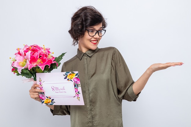 Woman with short hair holding greeting card and bouquet of flowers looking aside at her arm presenting something with arm celebrating international women's day march 8 standing over white background