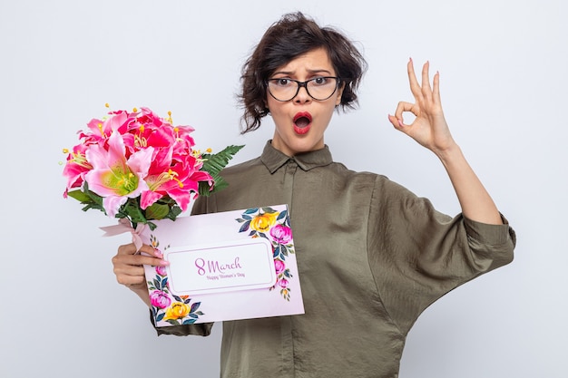 Woman with short hair holding greeting card and bouquet of flowers looking at camera confused and surprised doing ok sign celebrating international women's day march 8 standing over white background
