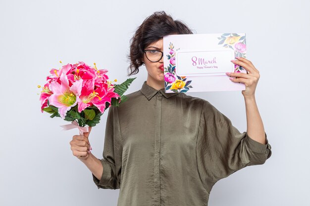 Женщина с короткими волосами, держащая поздравительную открытку и букет цветов, выглядит смущенной, празднуя международный женский день 8 марта