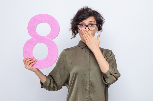 Free photo woman with short hair holding number eight made from cardboard looking at camera being shocked covering mouth with hand celebrating international women's day march 8 standing over white background