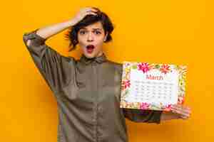 Бесплатное фото Женщина с короткими волосами, держащая бумажный календарь месяца марта, глядя в камеру, изумилась и удивилась празднованию международного женского дня 8 марта, стоя на оранжевом фоне