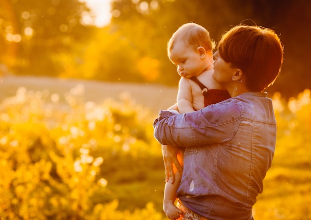 短い髪の女は夕方に芝生に立っている彼女の子供にキス