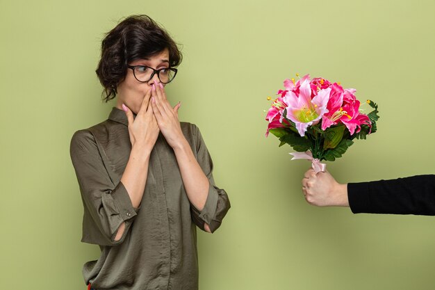 緑の背景の上に立って国際女性の日3月8日を祝う彼女のボーイフレンドから花の花束を受け取っている間驚いて幸せそうに見える短い髪の女性