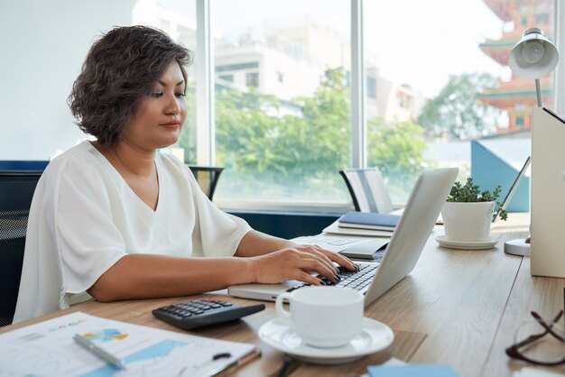 Woman with short wavy hair sitting at desk in office and working on laptop