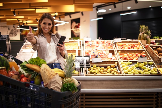 Woman with smart phone in supermarket standing by the shelves full of fruit at grocery store holding thumbs up