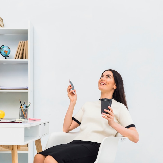Женщина с смартфон и чашка кофе, смеясь в офисе