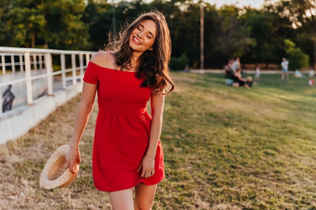 woman with straw hat in hand standing on the grass. Outdoor photo of fashionable brunette lady in red dress smiling at field.