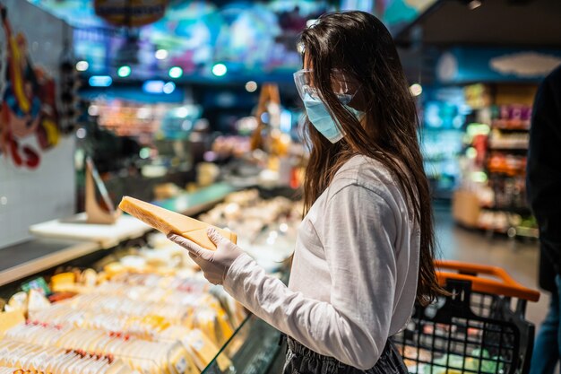 Женщина в хирургической маске и перчатках делает покупки в супермаркете после пандемии коронавируса. Девушка в хирургической маске собирается купить сыр.