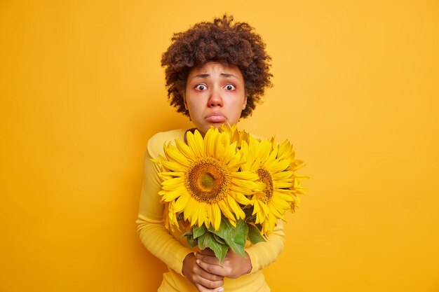 woman with symptoms of allergy has red swollen eyes holds bouquet of sunflowers isolated on vivid yellow 