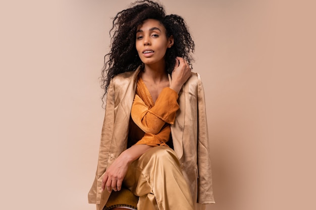 Woman with tan skin with perfect curly hairs in elegant orange blouse and silk pants sitting on vintage chair  beige wall.