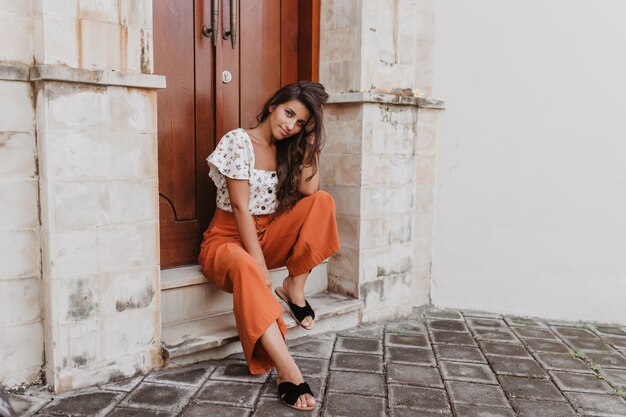 Woman with tanned skin in bright summer outfit posing sitting on threshold of old building with beautiful wooden door