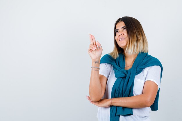 Woman with tied sweater showing gun gesture, looking up in white t-shirt and looking cheery. front view.