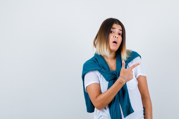 Woman with tied sweater in white t-shirt pointing at upper right corner and looking surprised , front view.