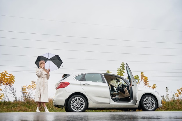 Woman with umbrella standing near car and speaking on phone