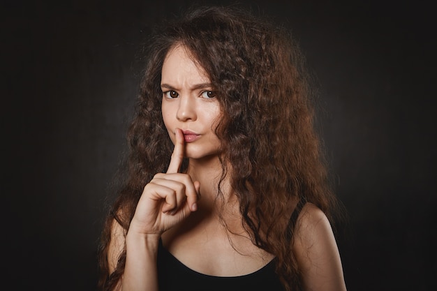Free photo woman with voluminous hair and clean skin frowning gesturing with fore finger at her lips, asking not to make nose while she's studying