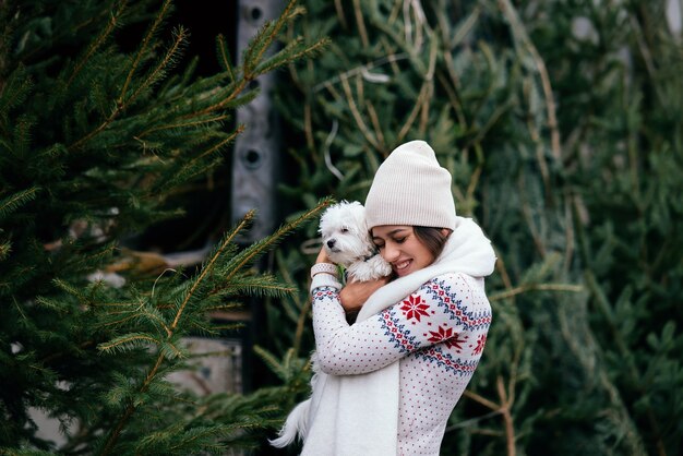 Woman with a white dog in her arms near a green Christmas trees at the market