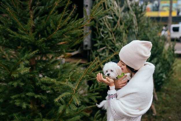 市場で緑のクリスマスツリーの近くで彼女の腕に白い犬を持つ女性