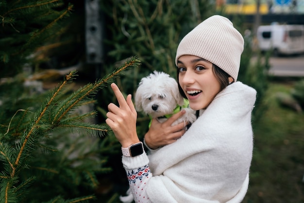 Woman with a white dog in her arms near a green Christmas trees