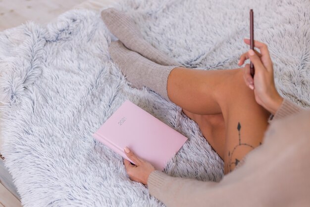 Woman in wool sock and sweater with pink notebook sign 2021, large tattoo on hip. Female sit on bed at home in bedroom.