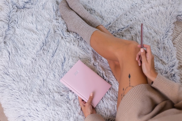 Woman in wool sock and sweater with pink notebook sign 2021, large tattoo on hip. Female sit on bed at home in bedroom.