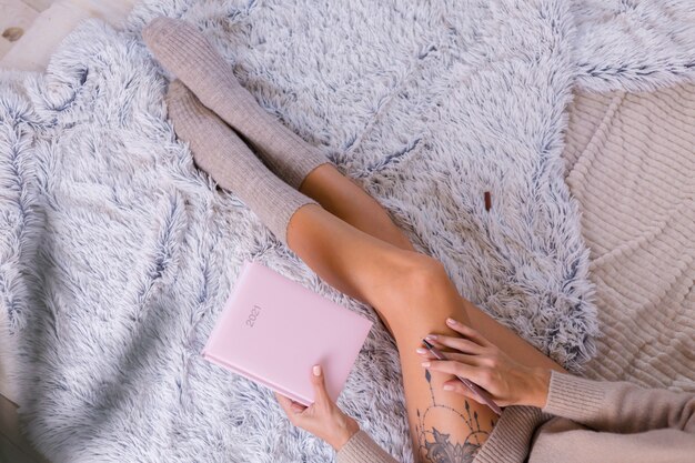 Woman in wool sock and sweater with pink notebook sign 2021, large tattoo on hip. Female sit on bed at home in bedroom.