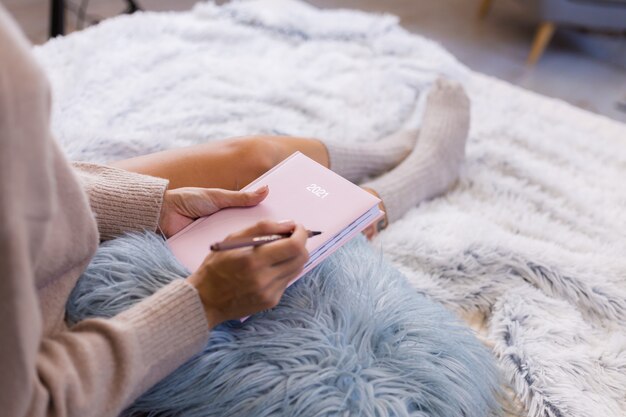 Woman in wool sock and sweater with pink notebook sign 2021, large tattoo on hip. Female sit on bed at home in bedroom.