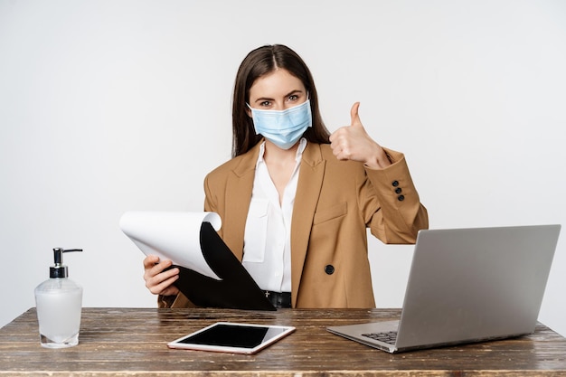 Free photo woman working in office at table holding clipboard with papers showing thumbs up wearing medical fac...