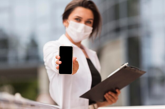 Woman working during pandemic outdoors showing smartphone while holding notepad