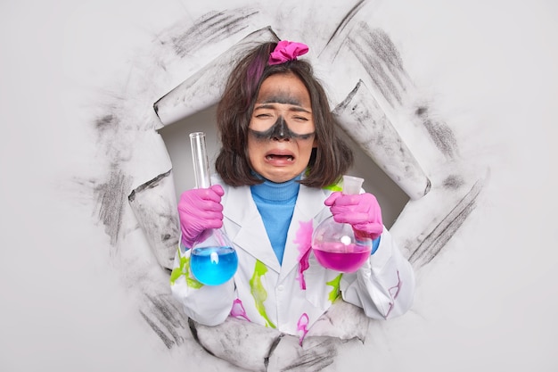 Free photo woman works as chemist holds test glass flasks with colorful solitude upset with results of experiment wears medical coat breaks through paper