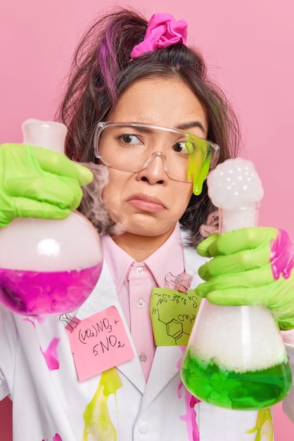 woman works in scientific laboratory holds two glass flasks with colored liquid bubbles and steam wears transparent glasses and medical coat on pink