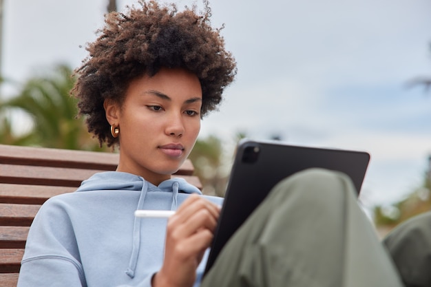 woman works on tablet does graphic design draws sketch works as freelance illustrator dressed casually poses outdoor uses electronic pen for writing