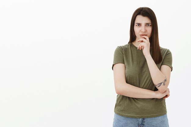 Woman worried and hesitant as hearing something disturbing thinking frowning from worry and intense disapproval holding hand on lip, considering what do, standing thoughtful and unsure over gray wall