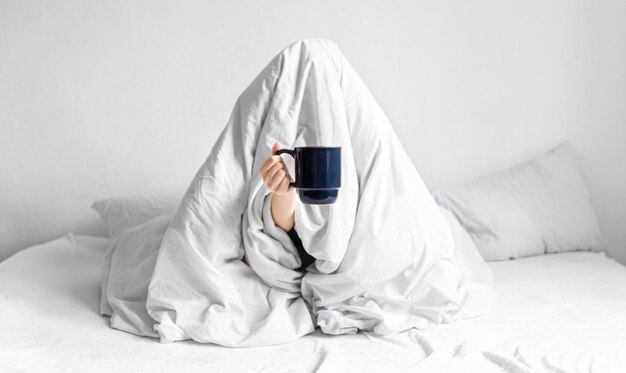 A woman wrapped in a blanket sits in bed with a cup of coffee in her hands