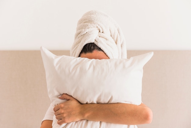 Woman wrapping her head with towel holding white pillow in front of her face
