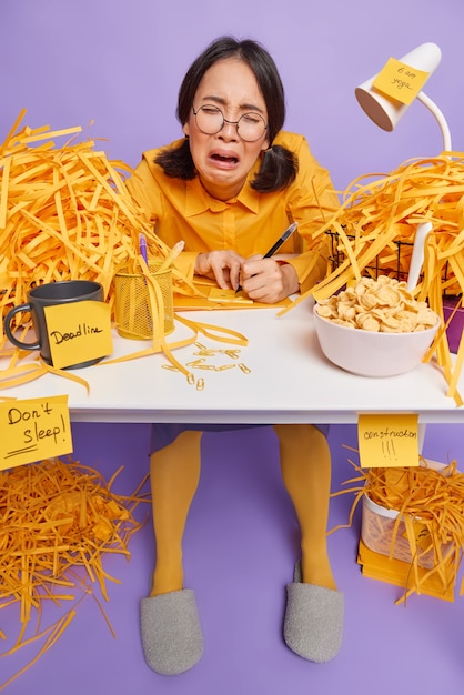 woman writes notes with pen feels very upset poses in coworking space with rubbish around feels tired of work on purple