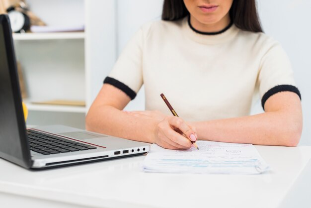 Woman writing on papers at table with laptop