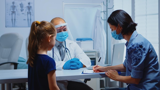 Woman writing prescription on clipboard listening doctor instructions. Pediatrician specialist in medicine with mask providing health care services, consultation, treatment in hospital during covid-19