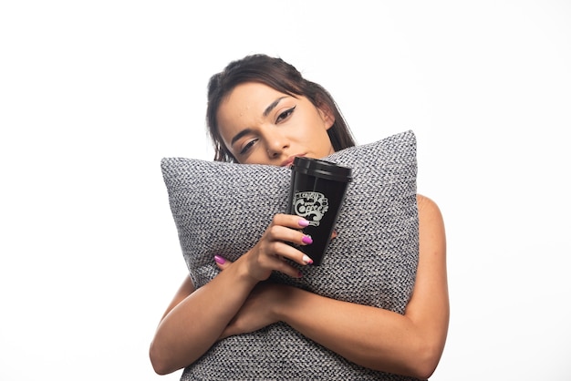 Woman yawning and holding a pillow with a cup on white wall