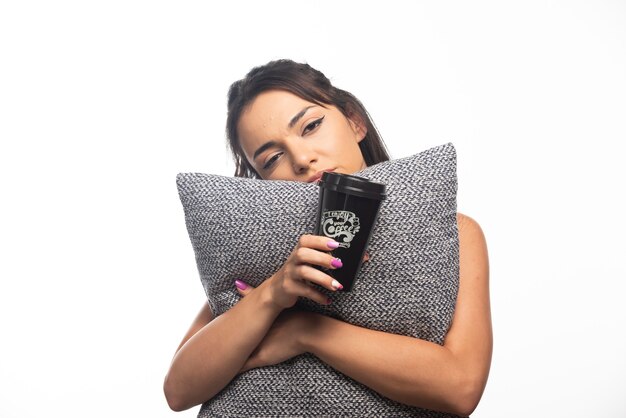 Woman yawning and holding a pillow with a cup on white wall