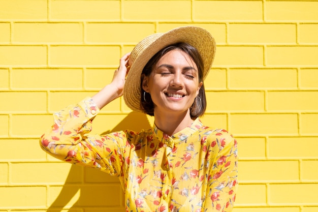 Woman in yellow summer dress and hat on yellow brick wall calm and positive, enjoys sunny summer days