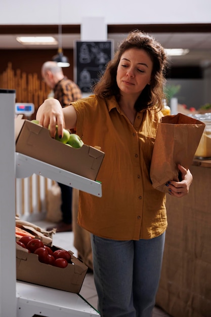 Free photo woman in zero waste shop purchasing farm grown vegetables, picking ripe green apples. client in plastic free local grocery shop using decomposable paper bag to save planet