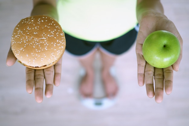 女性​は​体重計​で​計量し​、​リンゴ​と​ハンバーガー​を​保持しています​。​健康​に​良くない​ジャンク​フード​と​ビタミン​c​を​多く​含む​果物​を​選択する​決定​は​、​体​に​良い​です​。​ダイエット​の​コンセプト
