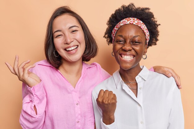 women celebrate achievement feel very positive smile broadly stand closely to each other dressed in shirts on beige