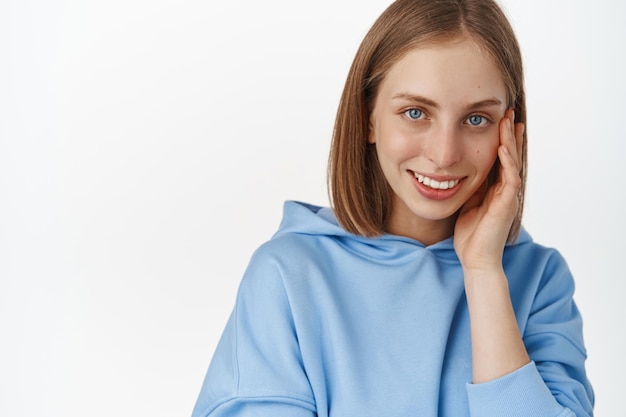 Women face and skin care. Portrait of beautiful blond girl touching her cheek, smiling and gazing happy at camera, dont be shy drop your cosmetics brand name, white background.