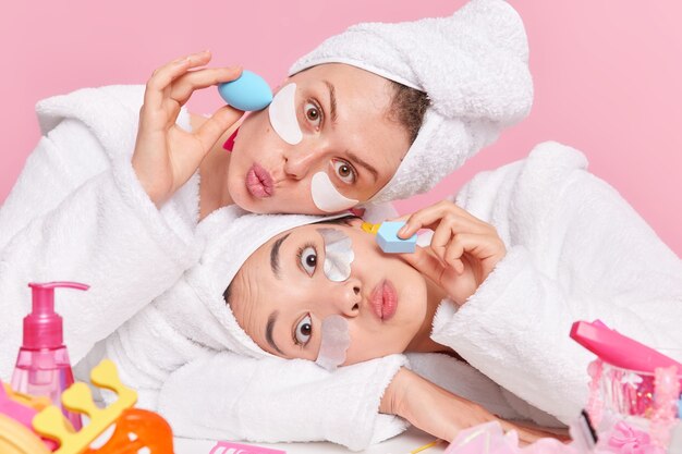 women have surprised face expressions keep lips folded lean heads on each other apply beauty patches foundation with sponges dressed in white bathrobes